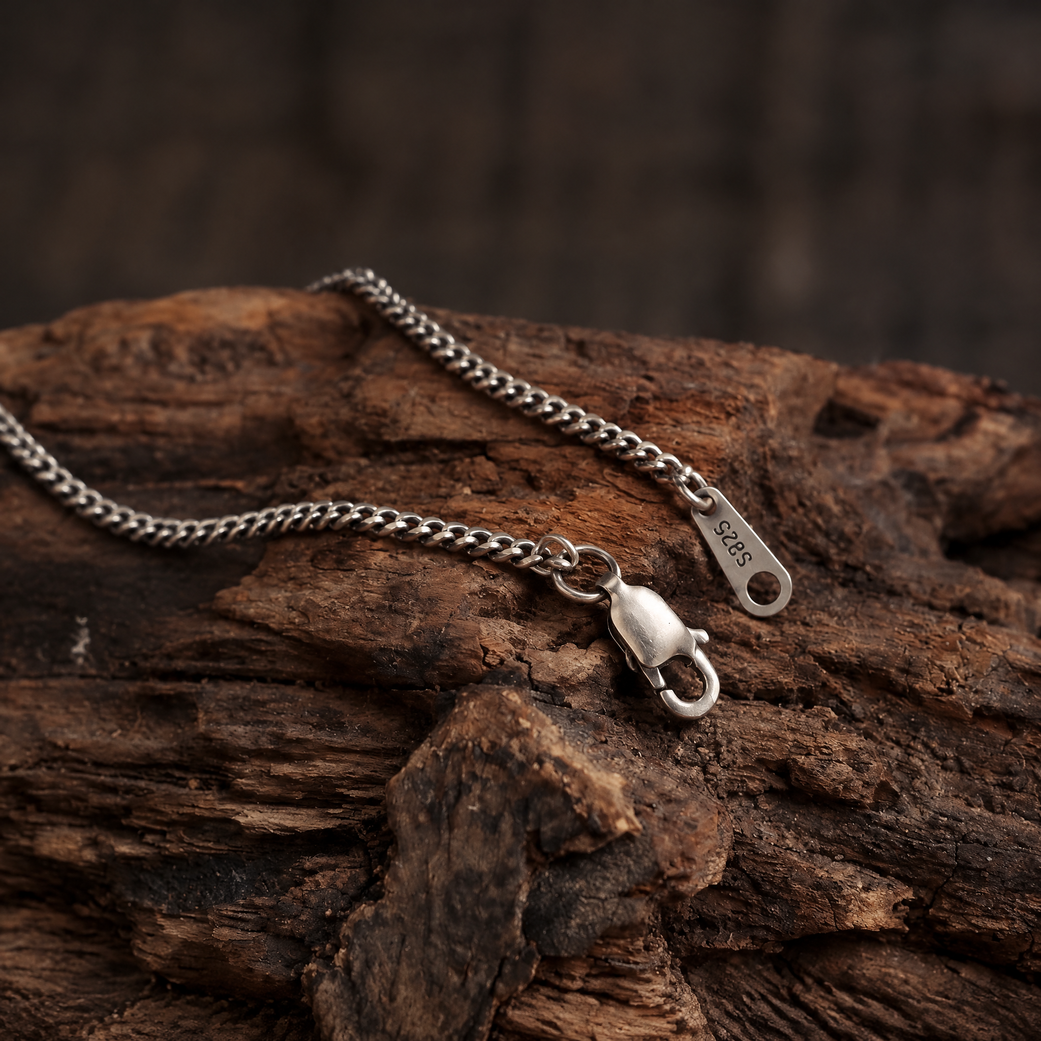 Close-up of an open lobster clasp and S925 tag on a Cuban link chain resting on rustic wood.